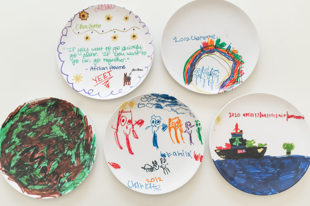 Five white dinner plates with colorful artwork drawn by kids are laid out, some show a battleship on an ocean, some have green and brown camouflage and others have African Proverbs drawn on them to show special birthday plates for children.