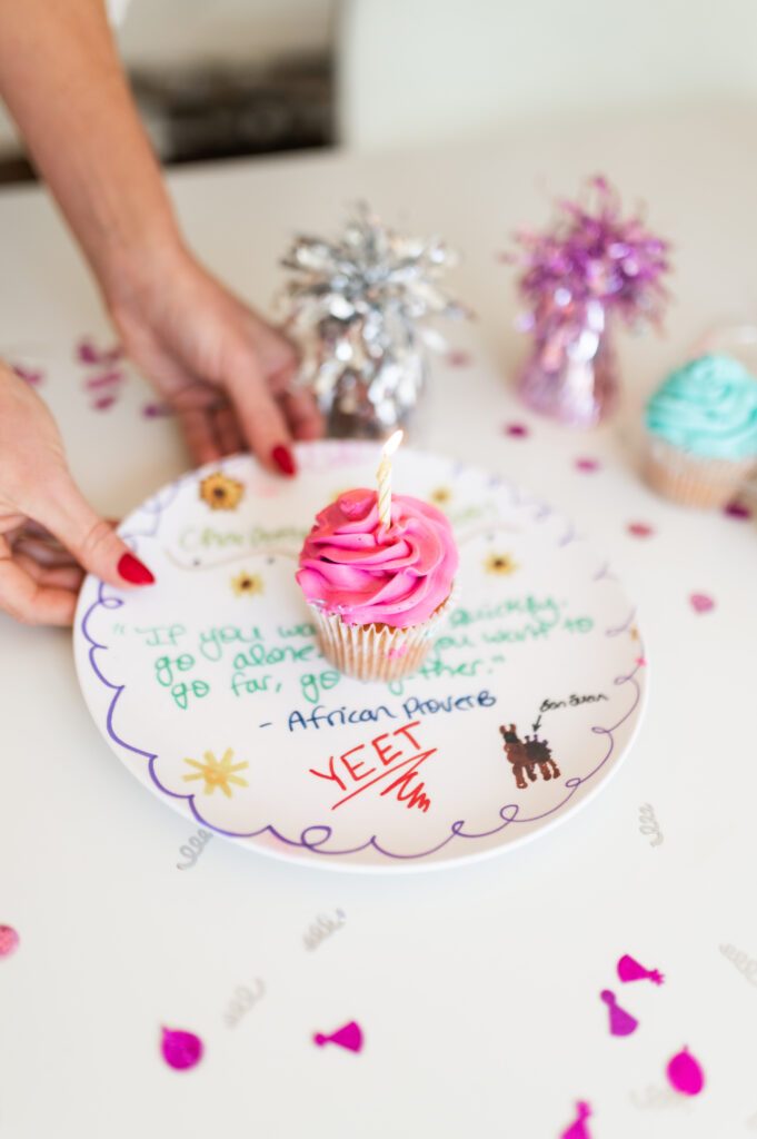 Dear Elly founder Kelly Page uses her hands to hold custom kids artwork keepsakes - this is a plate made by her daughter with an African Proverb quote. On it is a pink icing cupcake, and lots of confetti and party decorations are around it.