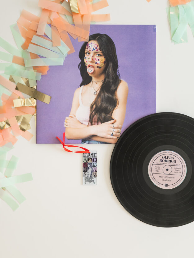Two fun concert ticket surprises for teens - shows the Olivia Rodrigo vinyl record album "Sour" laid out with a personalized vinyl record label showing the row, seat, and date of the concert I surprised my daughter with. It also shows an acrylic concert ticket ornament that has Taylor Swift Eras tour logo along with the date, city, row and seat for where we sat at the concert. Both are surrounded by orange and green garland as decoration.