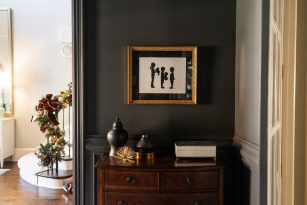 A custom child silhouette from photo is framed with black matting, gold frame and hangs in a home office with dark blue wall paint. Featured is also a piece of furniture and home accessories on it. You can see a foyer staircase banister in background where Christmas garland is hung.