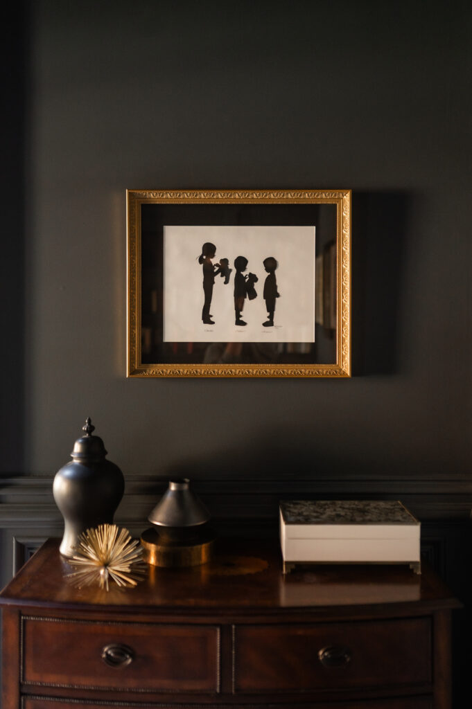 A framed personalized silhouette keepsake hangs in a home office. It has a rich gold frame, and hangs above a dark wood dresser with various accessories like vases and storage boxes above it.