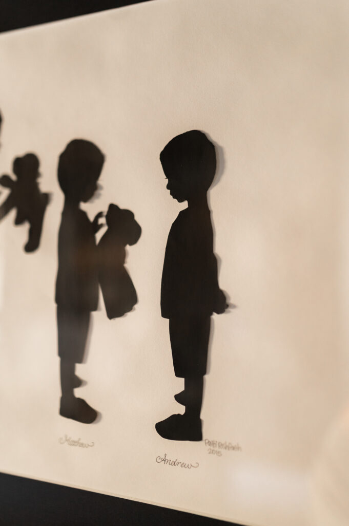 Sentimental wall art for parents has silhouette cut outs made by a woman owned small business called Heart, Paper Scissors. Two boys are cutout in silhouette. One is holding a stuffed animal, the other has his hands behind his back.