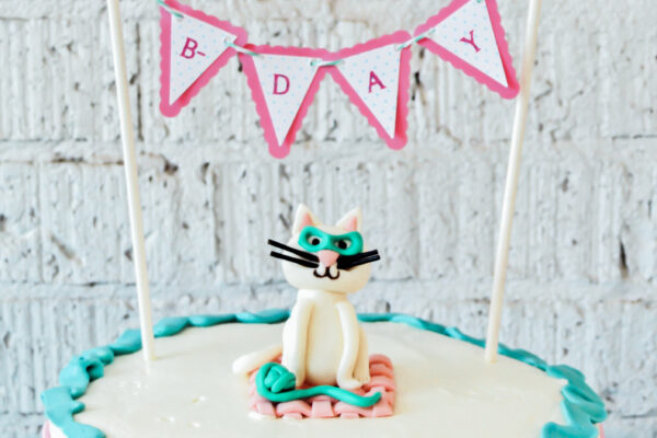 A birthday cake with a fondant cat and a cake topper that says "Happy Bday" on top of a teal and pink cake shows how to throw a kitty themed birthday party.